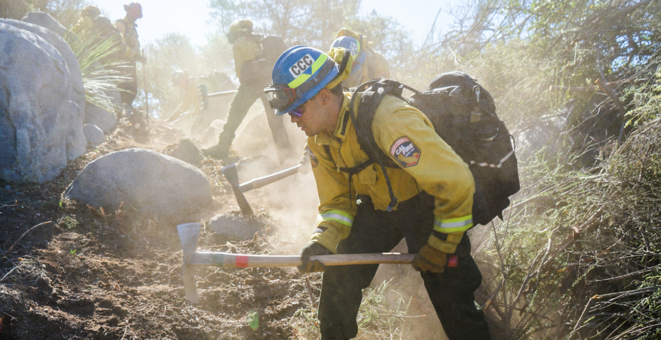 corpsmember in fire gear using pulaski to cut hand line on a dusty trail