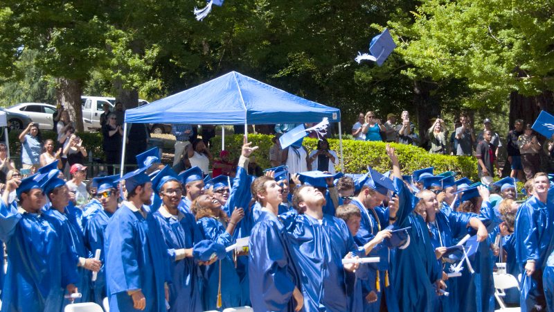 A large group of graduates in blue robes toss their caps into the air.