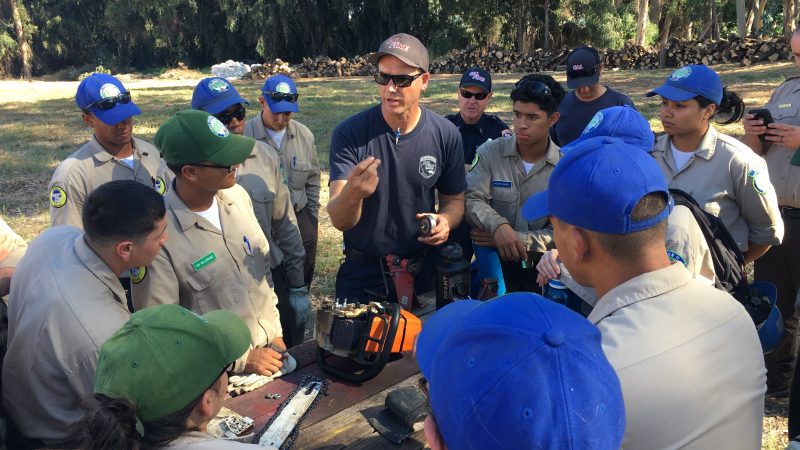 A group of Corpsmembers circle around a CAL Fire instructor, as a disassembled chainsaw lies on the table in the middle of the group.