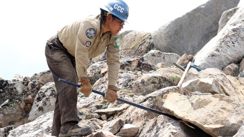 Female Corpsmember wearing PPE uses rock bar to lift giant rock during trail work in backcountry