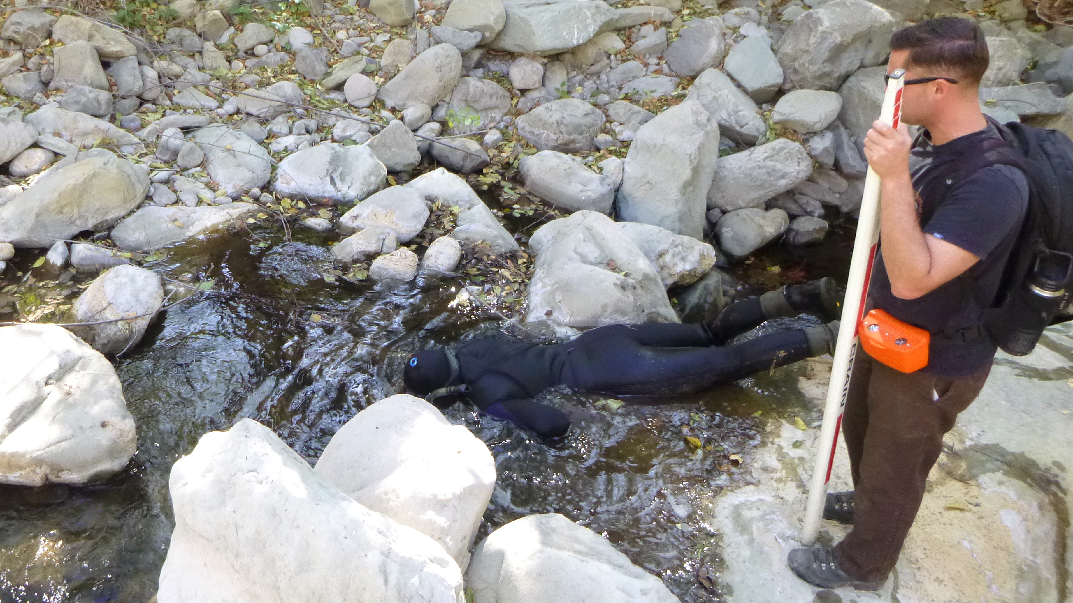 Corpsmember in snorkel gear peers into stream, while another Corpsmember watches over them on top of a nearby rock