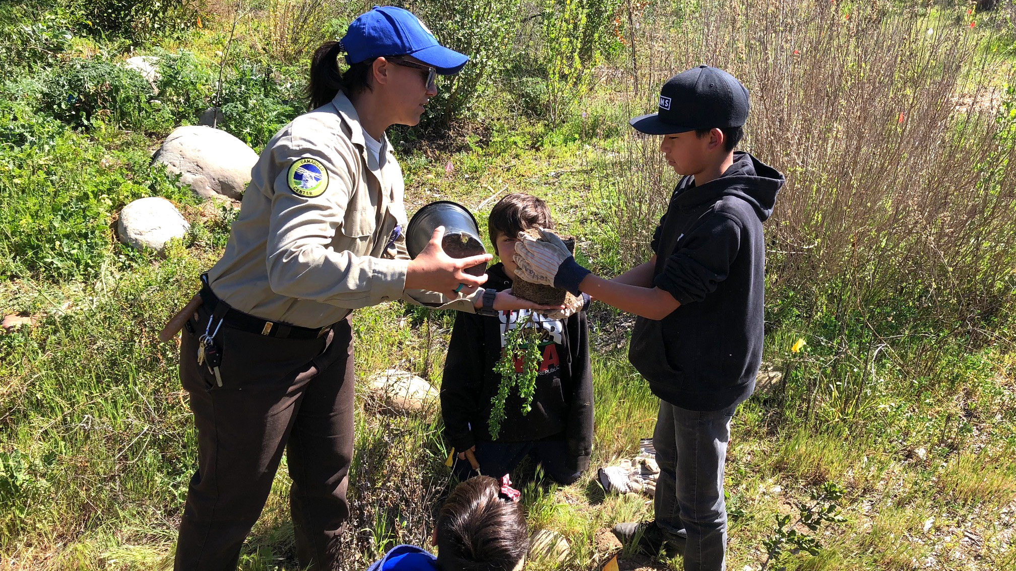 Female Corpsmember holds pot of plant while showing three young children how to properly plant it