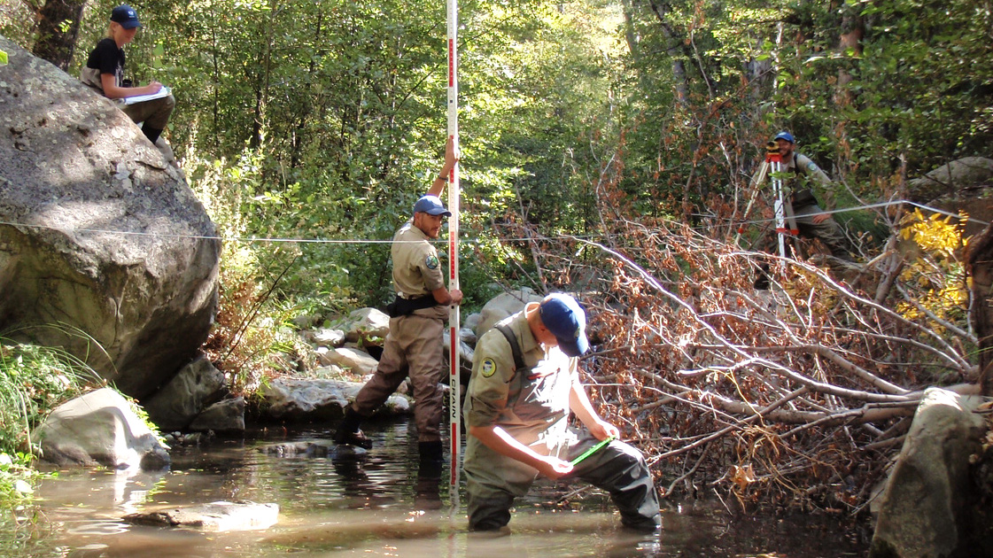 Two Corpsmembers in hip-waders stand in creek taking measurements, while others in background record data