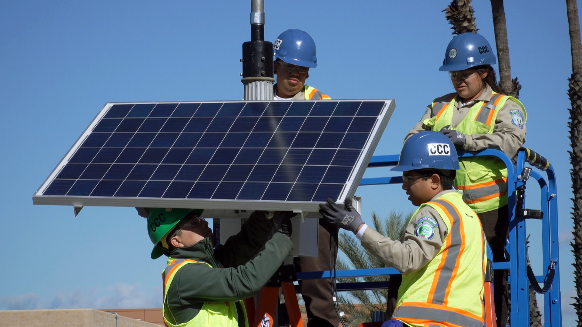 Image. Corpsmembers in protective gear afix a solar panel to a street light