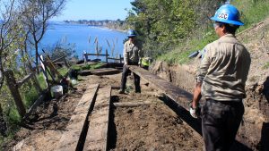 Two Corpsmembers, one in foreground and one in the background, work together to carry a large wooden plank.