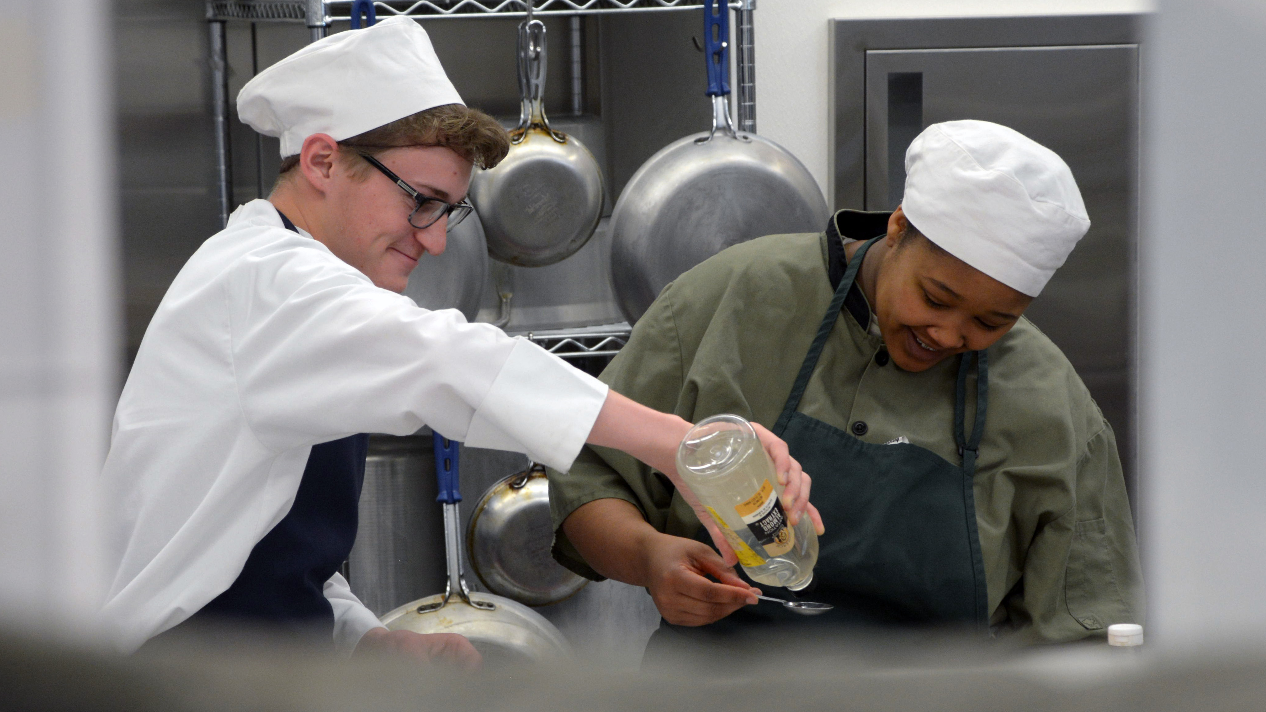 One culinary Corpsmember holds out a spoon as another pours liquid into it from a bottle.