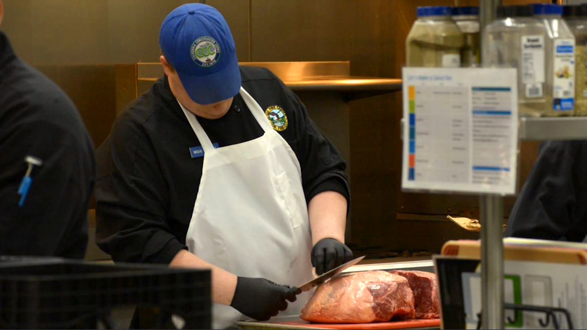 A Corspmember uses a knife to trim fat off of a beef roast.