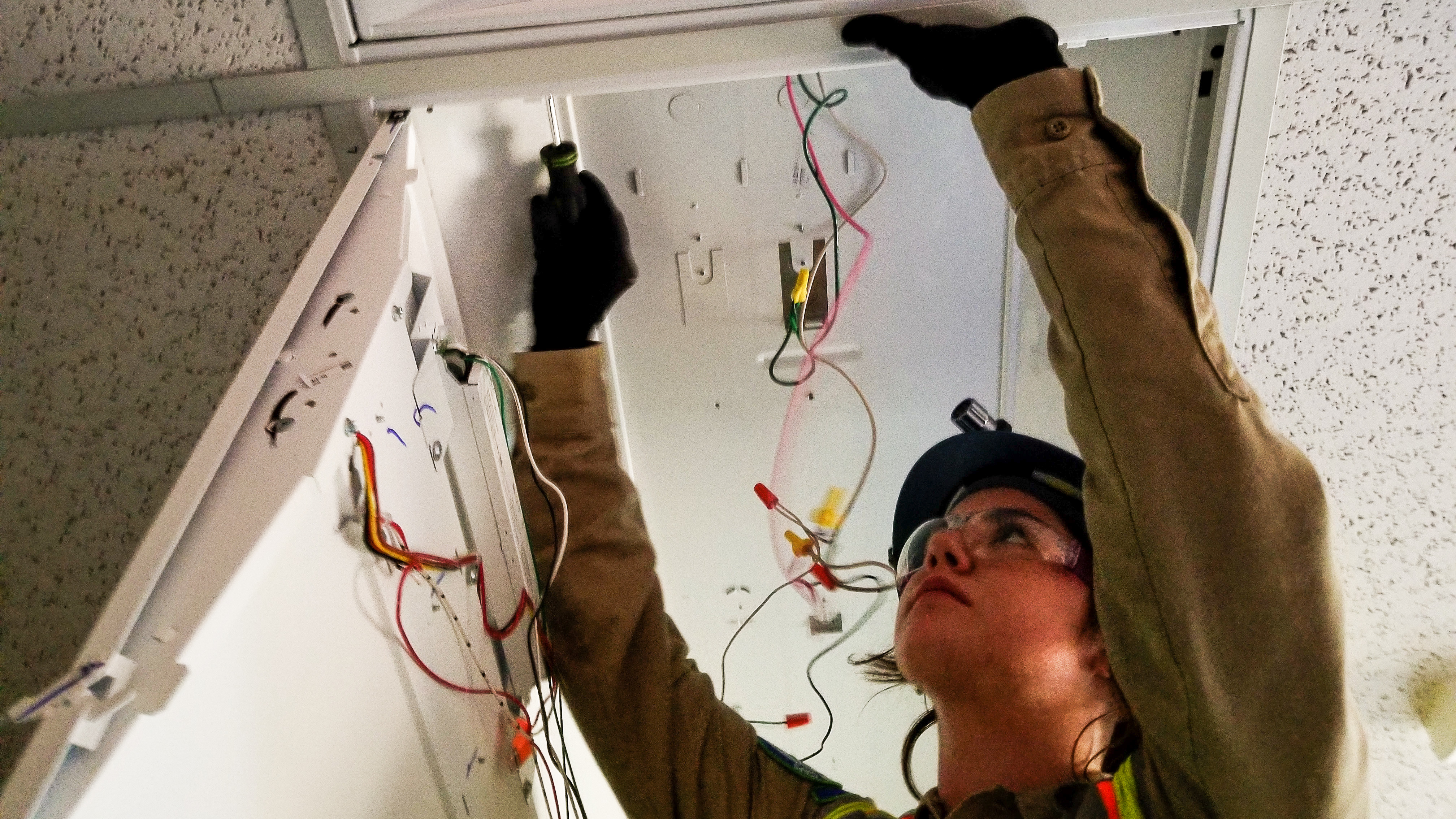 Image. Female Corpsmember in protectiv equipment looks at wires in overhead light fixture