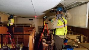 Three Corpsmembers in protective equipment stand atop ladders changing out light fixtures inside a court house clerk's office