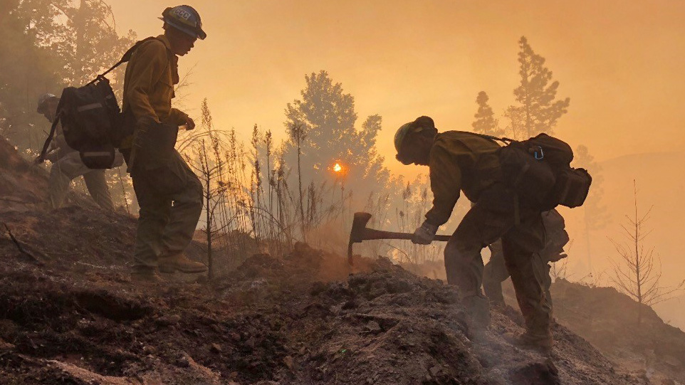 Two male Corpsmembers use pulaskis to cut fire line as sun sets among smoke