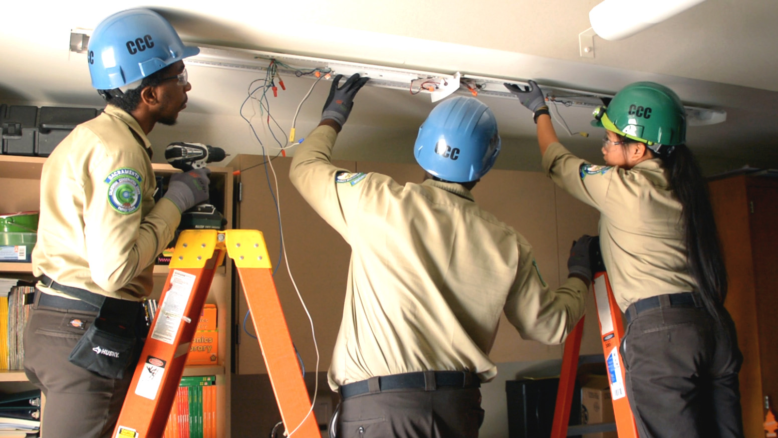 Image. Three Corpsmembers on ladders in protective equipment look at how to install energy efficient light fixture