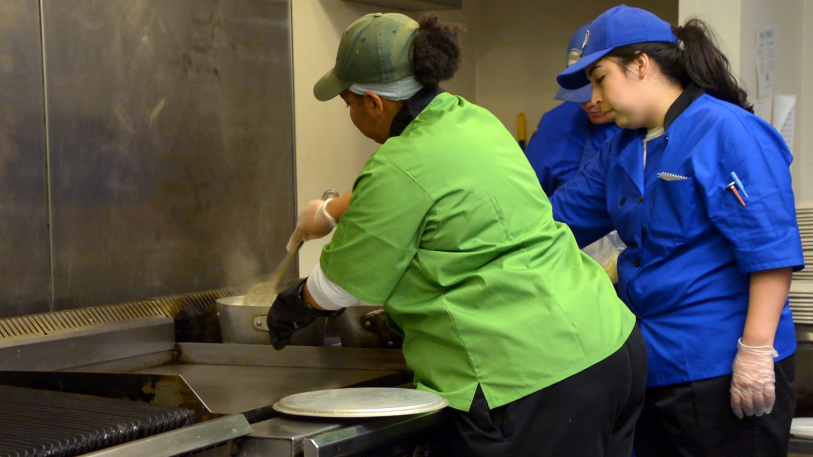 A green-hatted Corpsmember stirs a pot on the stove as two blue-hatted Corpsmembers observe.