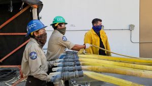 Corpsmembers in full uniform stand near water pipes where hoses are attached and look at the hoses.