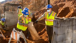 Corpsmembes wearing yellow safety jackets and blue helmets use shovels to fill sand bags.