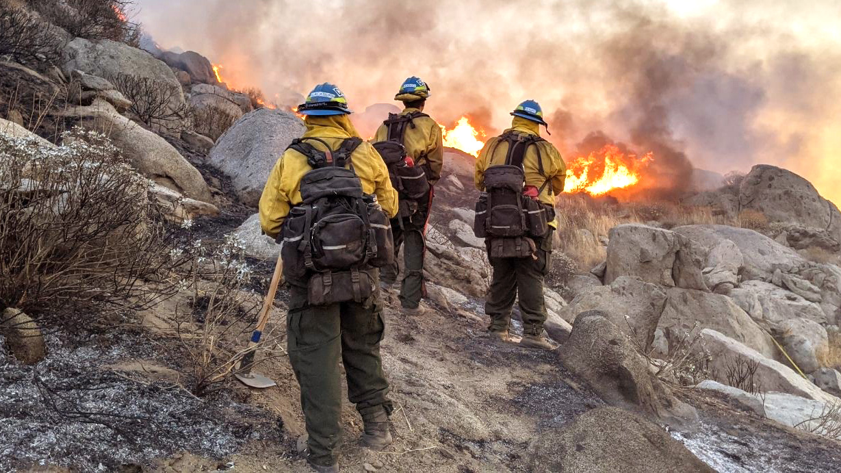 firefighters standing in rocky terrain with flames approaching