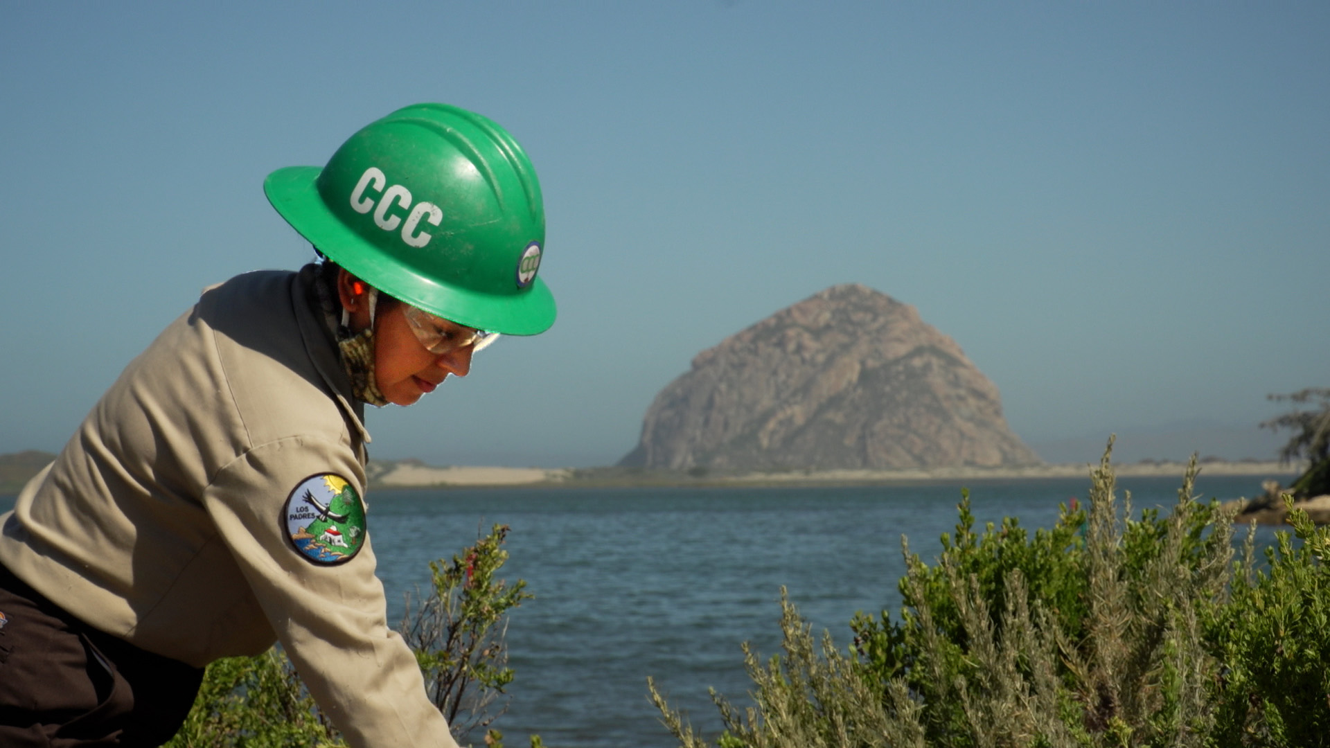 Female Corpsmember with green helmet ans safety glasses extends arm out of frame in foreground, Morro Rock and Morro Bay are in background