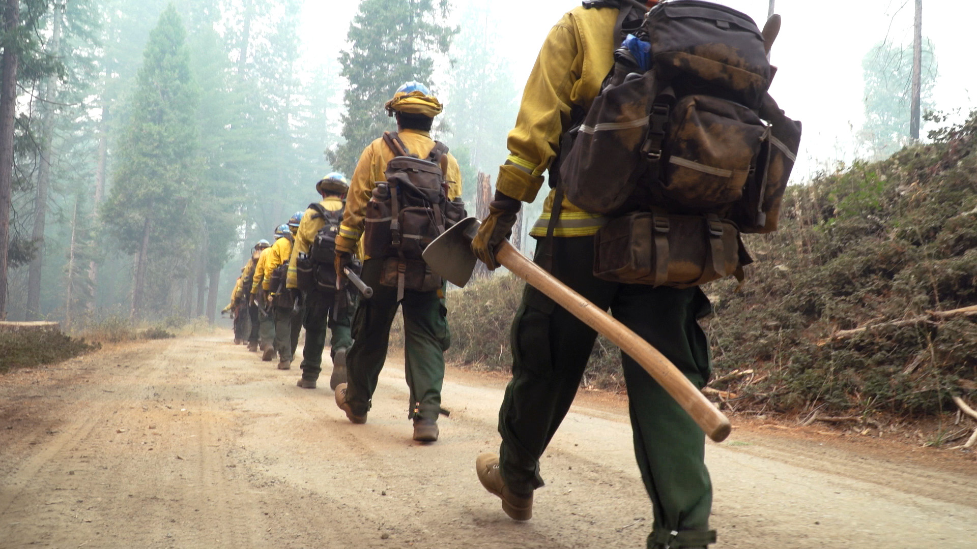 corpsmembers walking holding hand tools and wearing backpacks, walking on dirt road heading toward tall trees and smoke