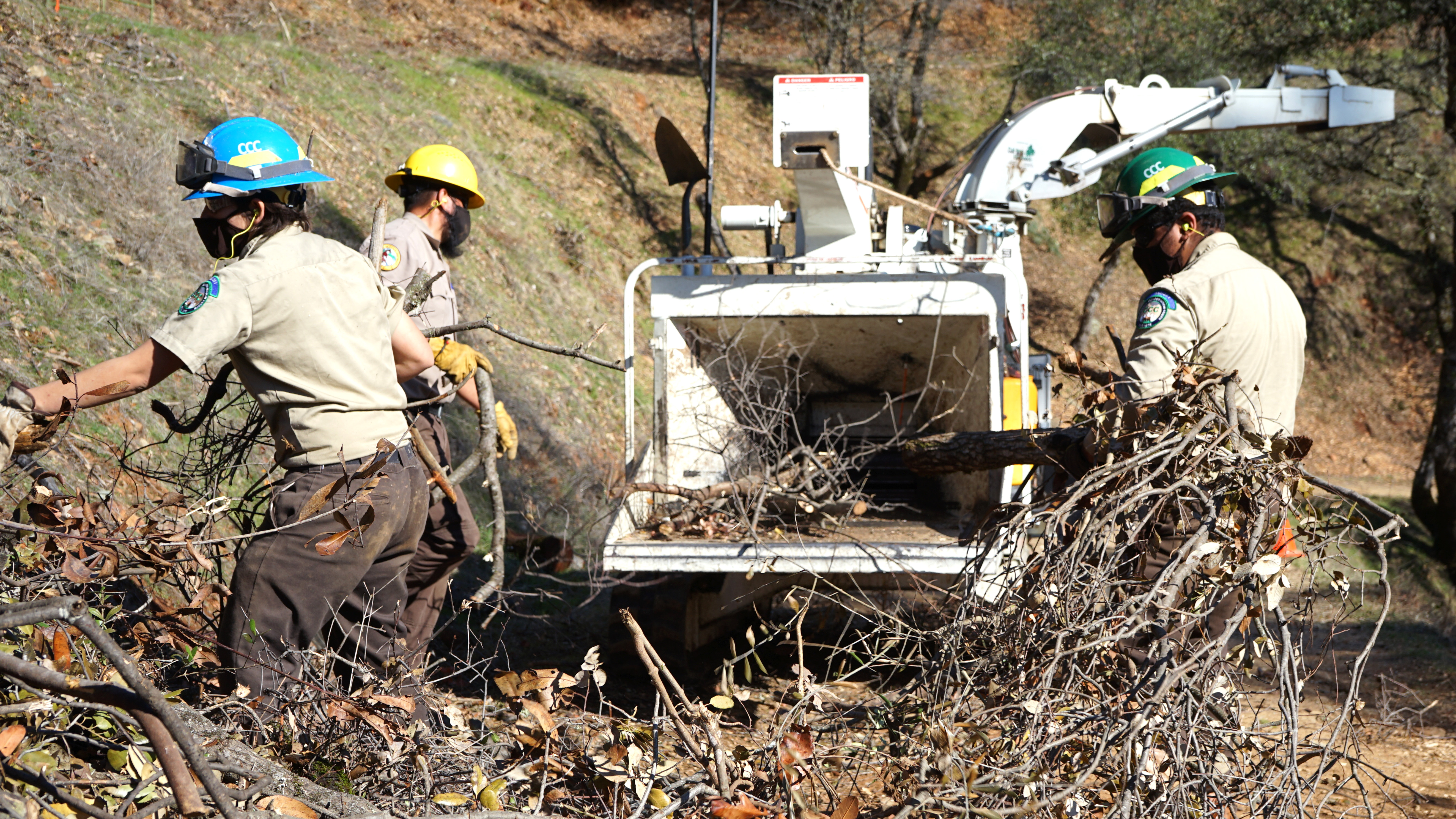 Corpsmembers pull branches from debris pile and haul them toward a wood chipper in the background of image