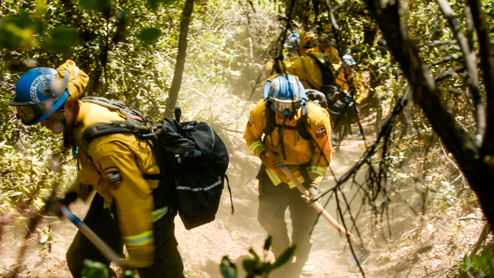 two corpsmembers in fire gear and with hand tools walk through wooded area, with tree limbs and leaves in foreground