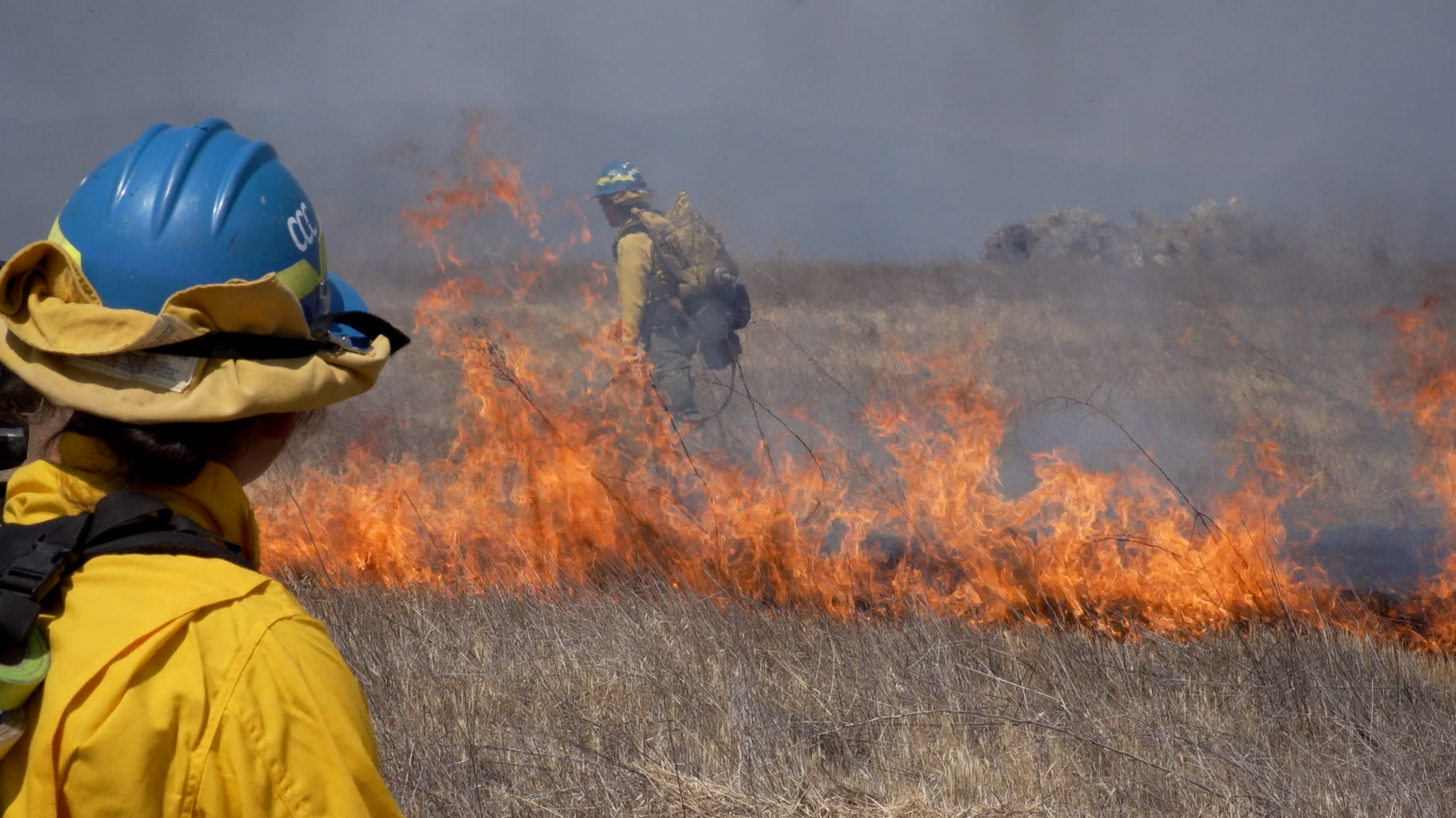 female in fire gear in foreground watches flames flicker in background