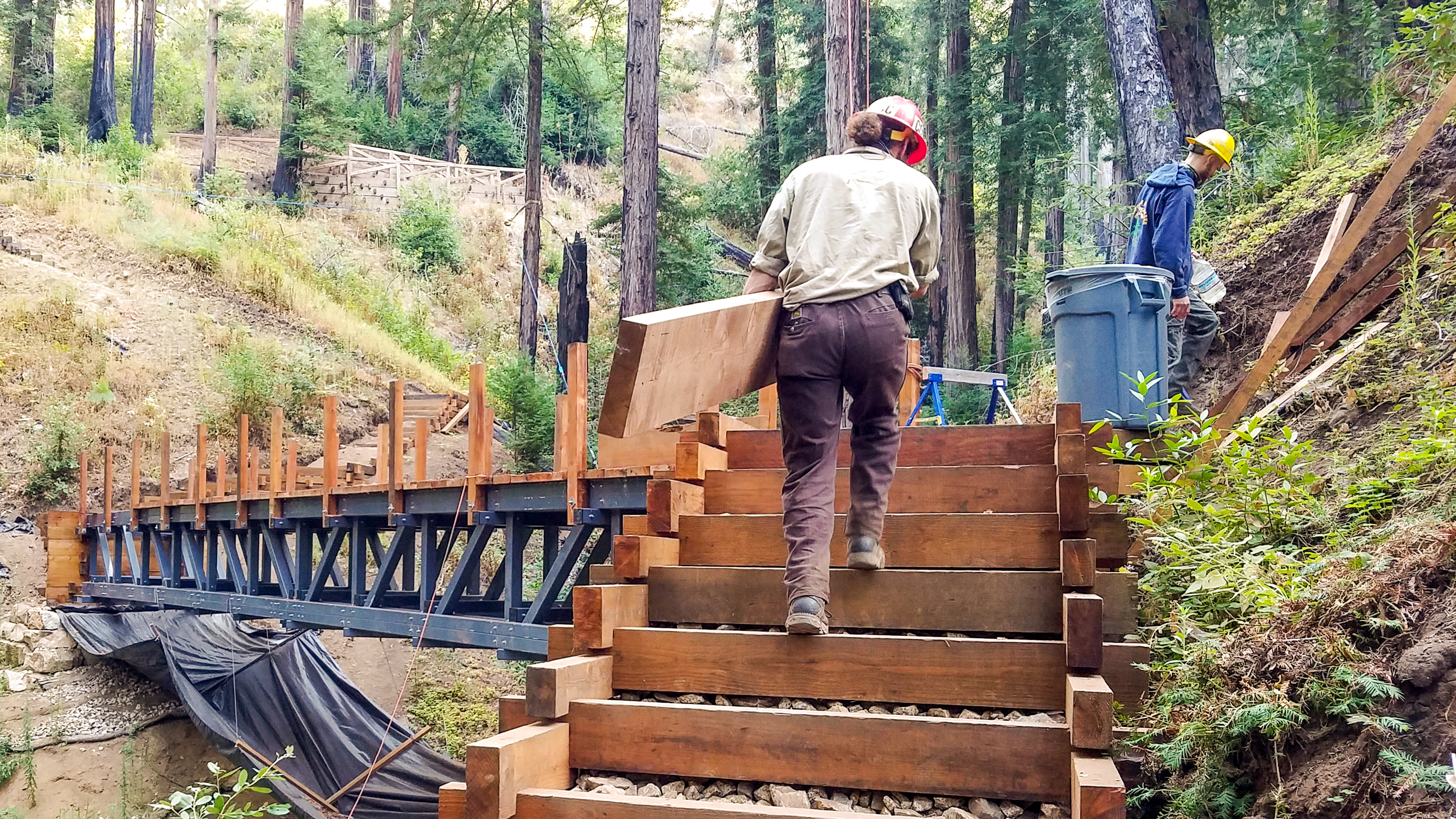 corpsmember carrying wood beam climbs stairs near bridge