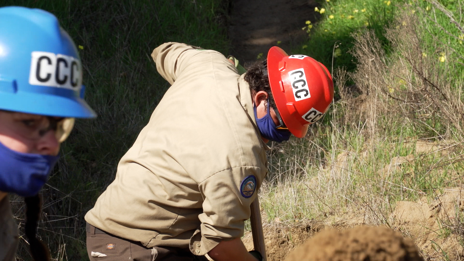 male corpsmember in red helmet head facing down as he uses a shovel