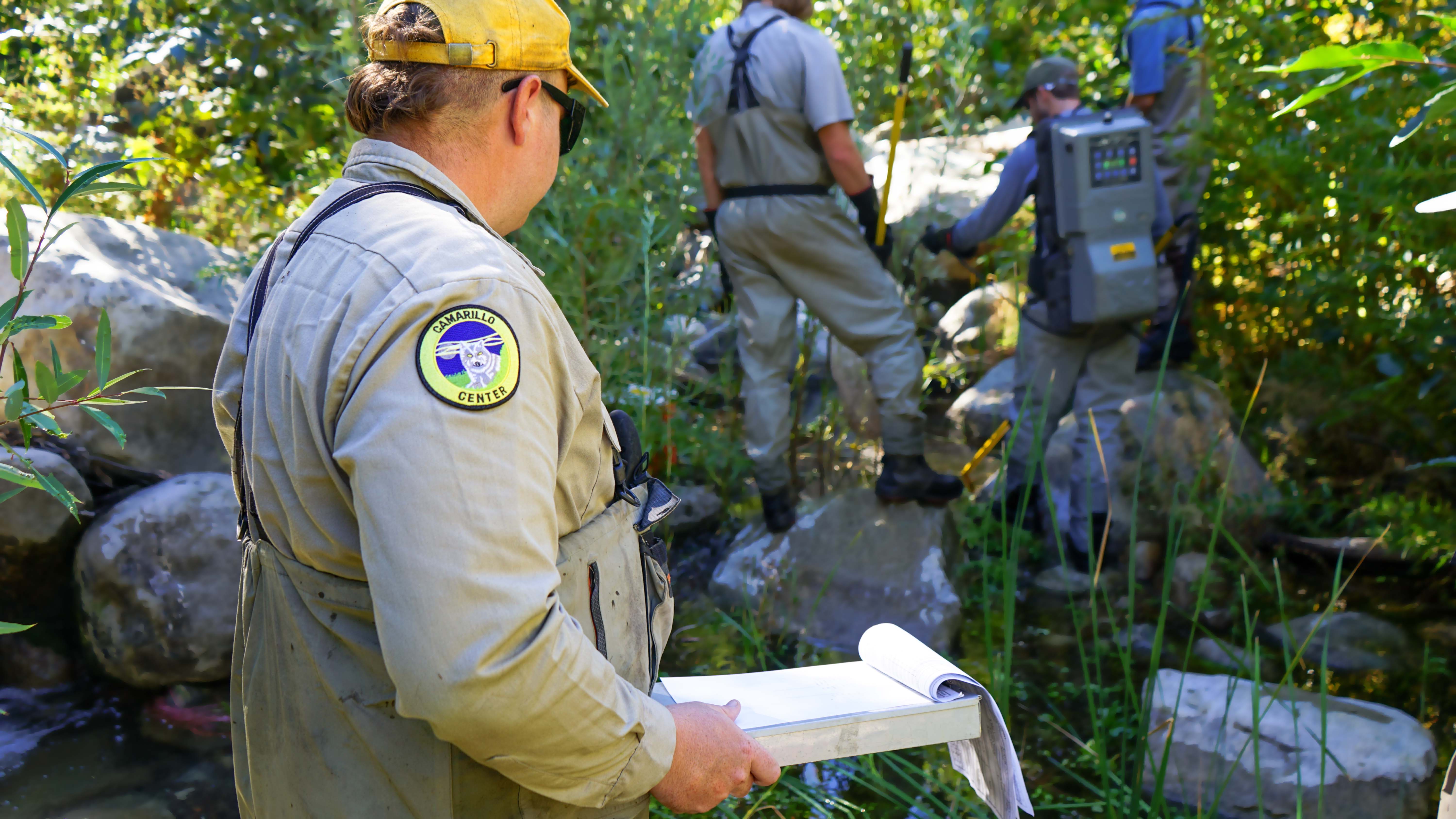 male in yellow hat and waders holds clipboard as others in distance relay information to him to record