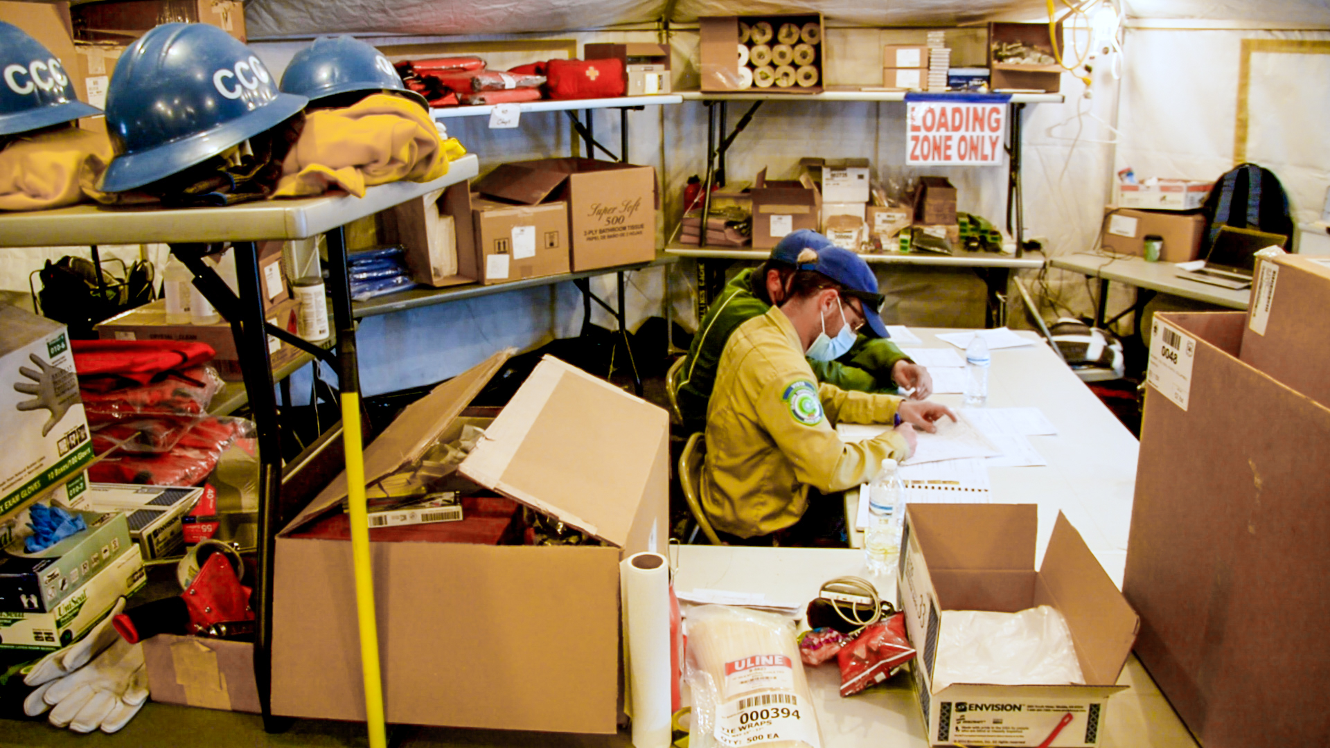 Two men sitting at table in supply tent surrounded by shelving and boces