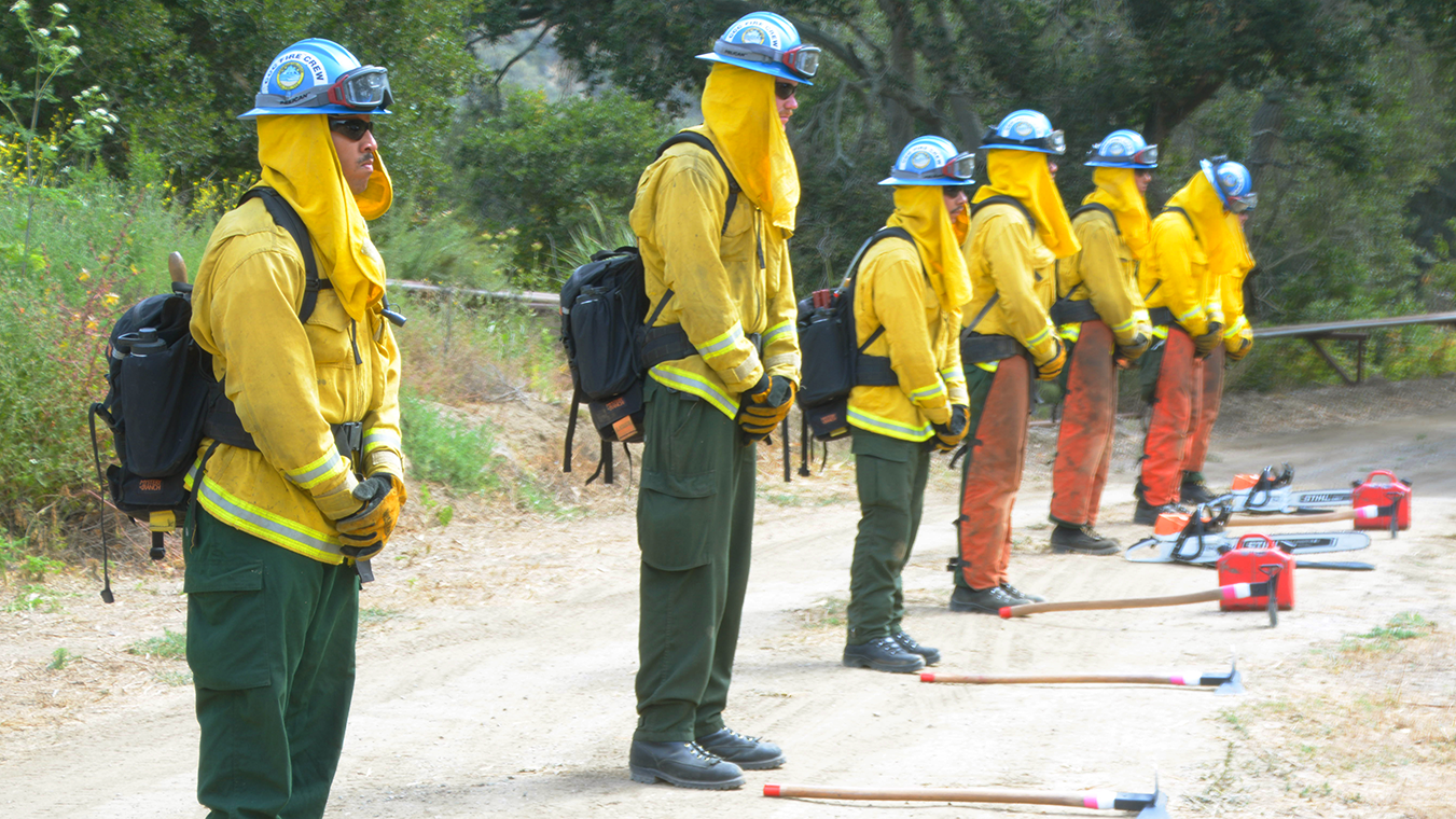 corpsmembers in firefighting nomex stand ready with tools at their feet