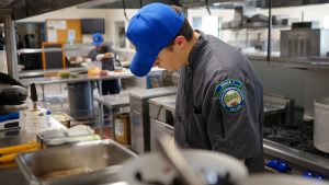 forks out of focus in the foreground, corpsmmeber in hat standing in kitchen