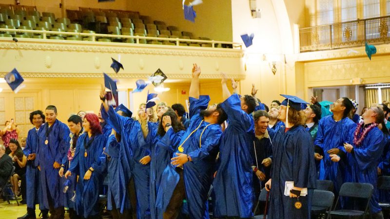 corpsmembers in graduation gowns tossing caps into the air