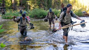 Corpsmembers cross a creek carrying tools.