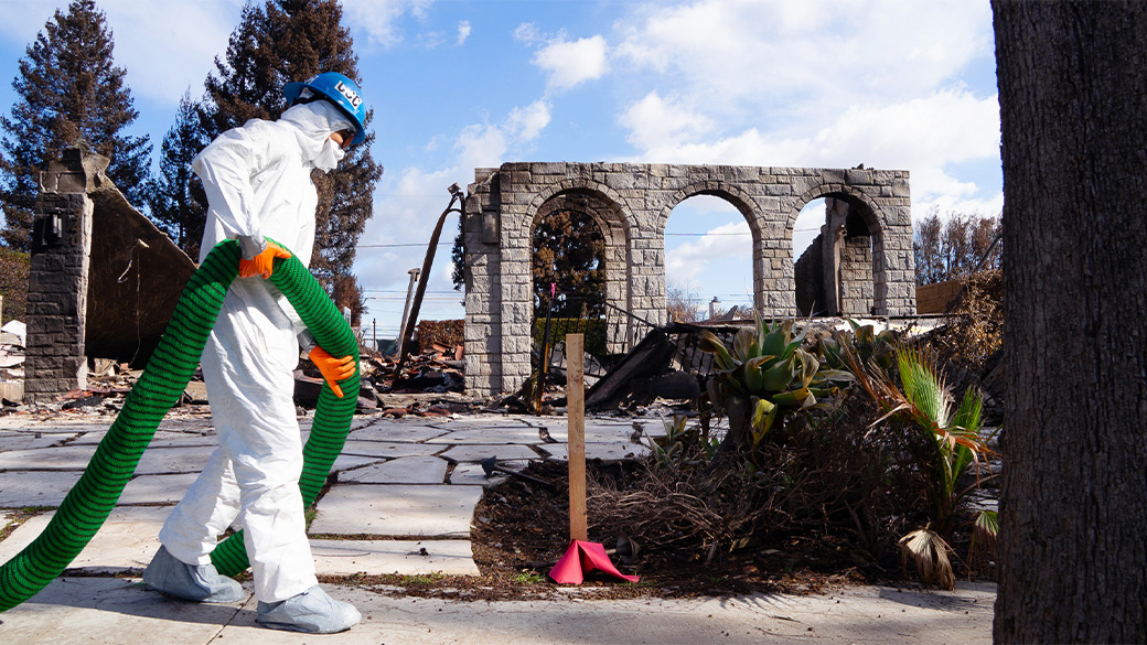 corpsmember in full tyvek holding a silt sock in front of a burned out hoe