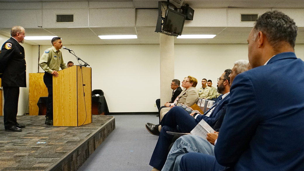 in foreground a man listens intently from a seated position, with a speaker at a podium in the deep background
