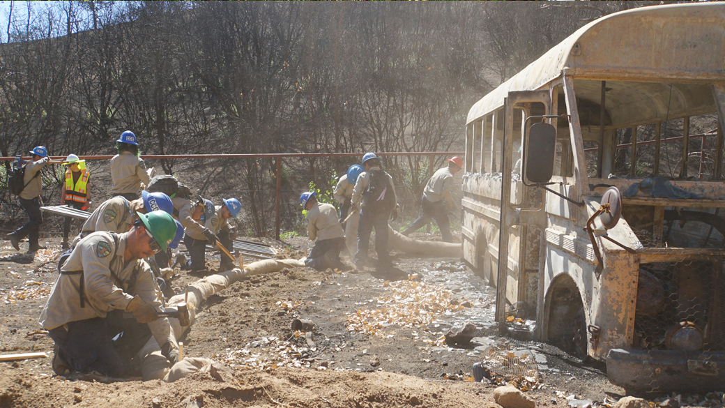 corpsmembers in helmets and uniforms hammer stakes into straw wattle surrounding a burned out bus