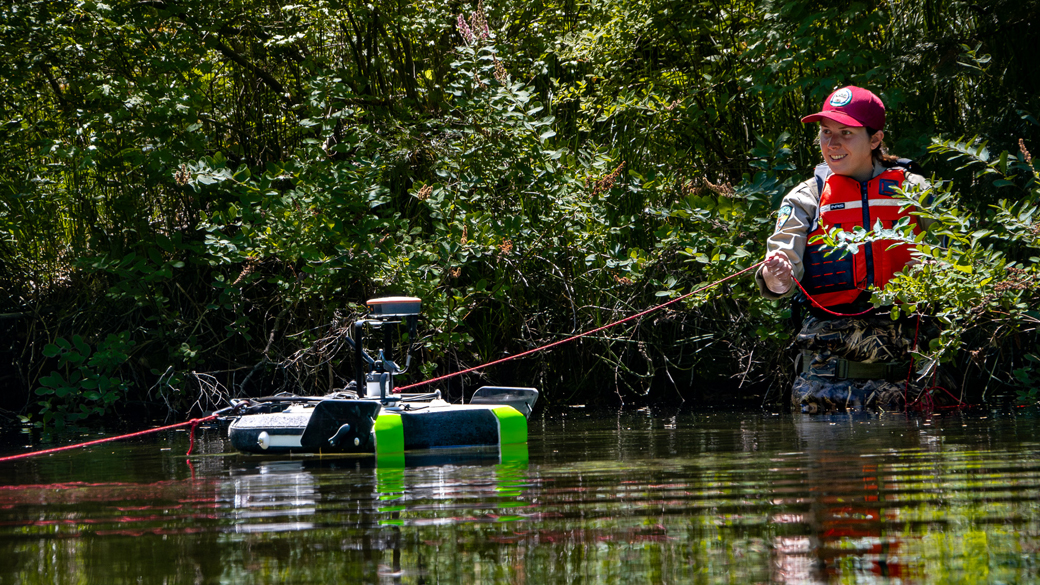 Interns_Chico_DWRBoogeyBoard_072325_1040x585 female standing in waders in creek pulling a rope with a boogey board on the creek