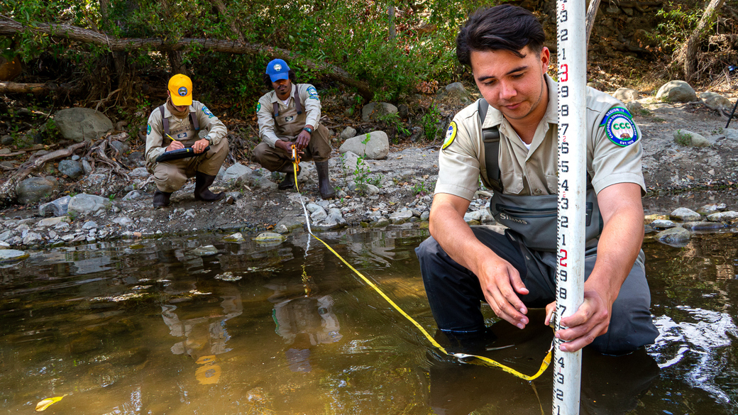 two men on bank of creek holding measuring tape and taking notes, third man in creek holding a measuring device in water