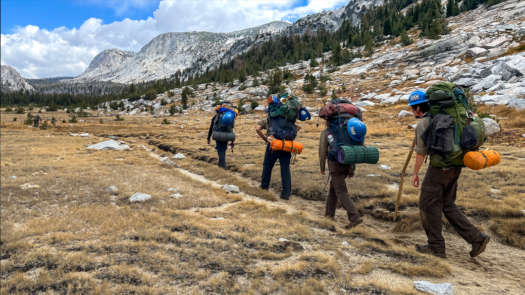 corpsmembers carrying backpacks hike through a remote stretch of wilderness with snow on the hillsides behind them