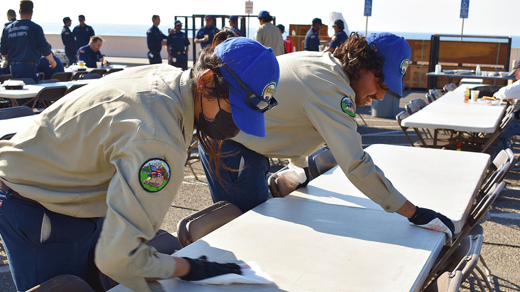 two corpsmembers with gloves on wipe down a dining table