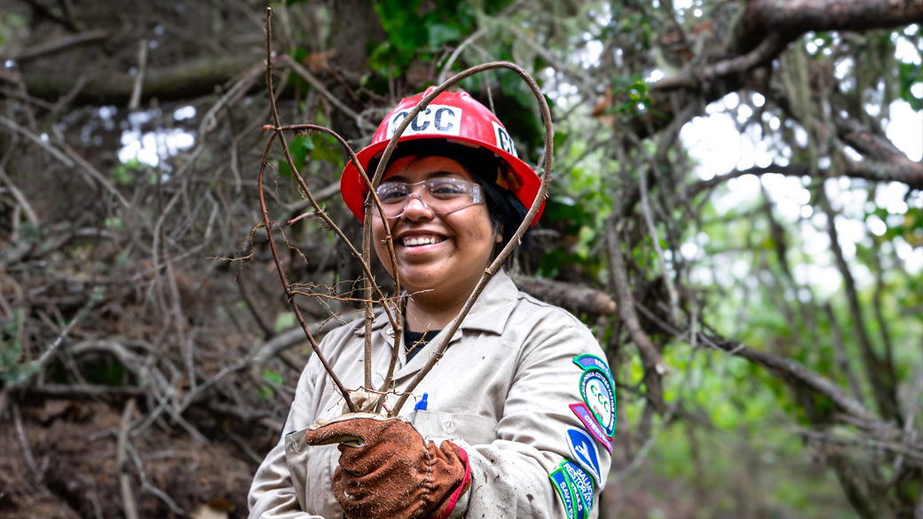 Fortuna_MalelDunes_HeartOfIvy_071024_1040x585 Female Corpsmember smiling while holding up English Ivy shaped in the form of a heart.