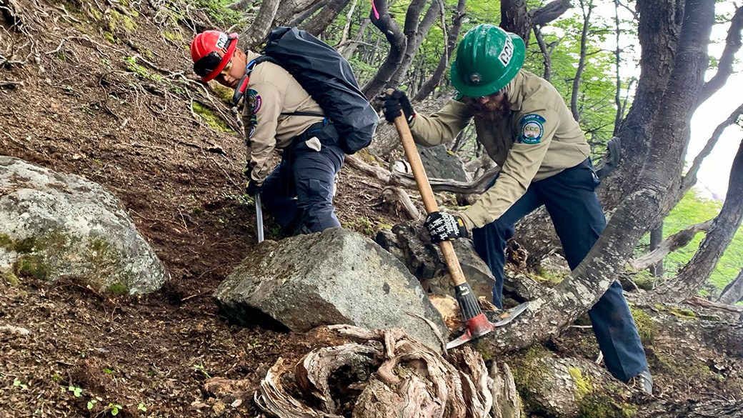 Corpsmembers using hand tools on a trail to move a large rock.