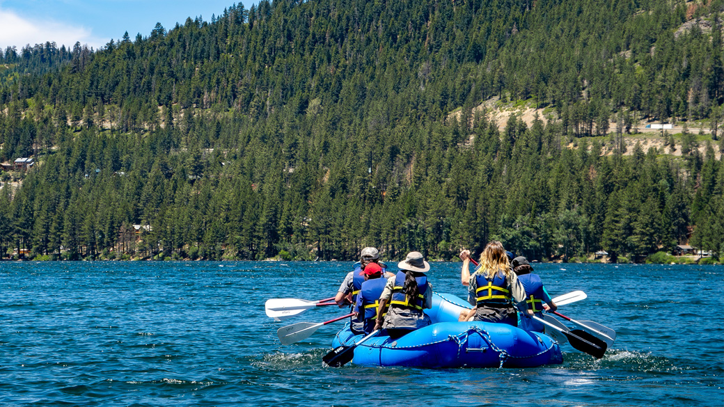 Internship_StateParks_DonnerRafting_062724_1040x585 A raft with people rowing it moves across Donner Lake with a forested mountain in the background.
