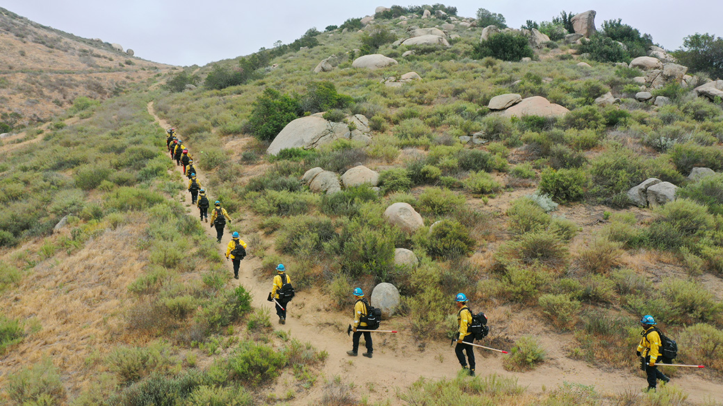 corpsmembers in fire equipment hiking up hill holding hand tools