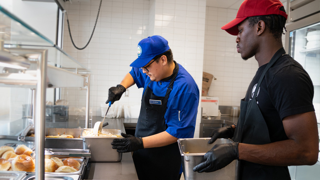 Special_Culinary_ServingTeamWork_111324_1040x585 corpsmember stirs in a serving tray with another corpsmember arriving next to him with another serving tray