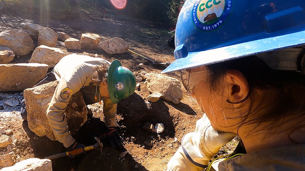 man watching other man hit chisel on rock