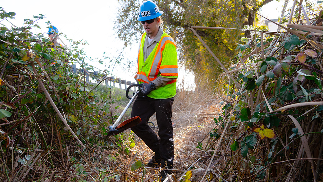 male corpsmember walking between debris piles in culvert while holding weeddwhacker
