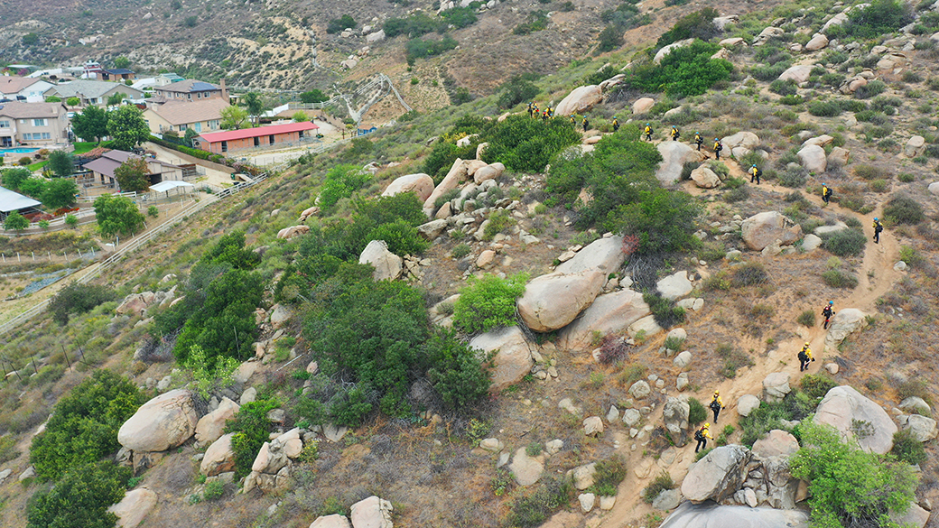 corpsmembers in fire gear, on the right, snake around a trail in steep terrain with homes nearby