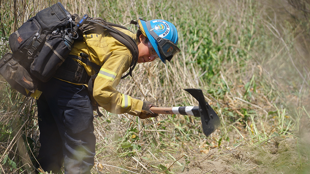 female corpsmember in fire gear bends over in dusty trail while swinging a rogue hoe