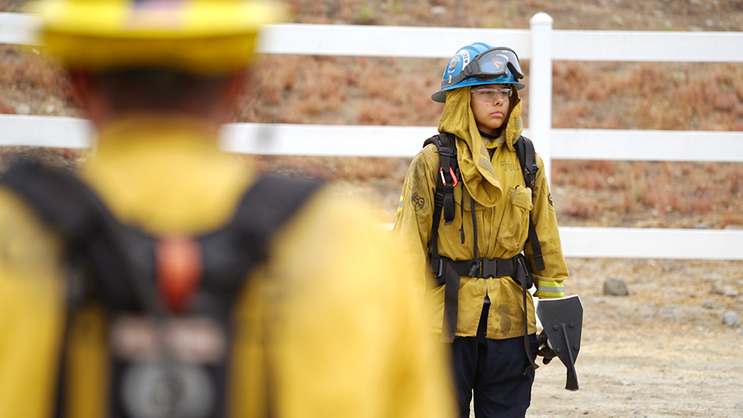 female corpsmember in fire gear holds rogue hoe during line out in background