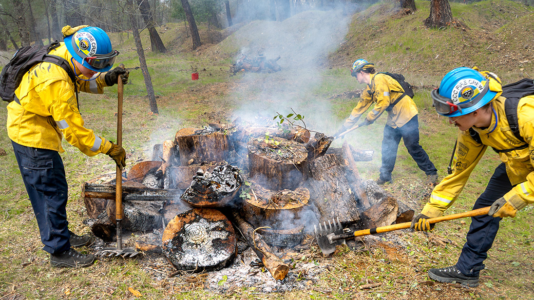 corpsmembers in fire gear using mcleods to stoke a burning wood pile smoldering
